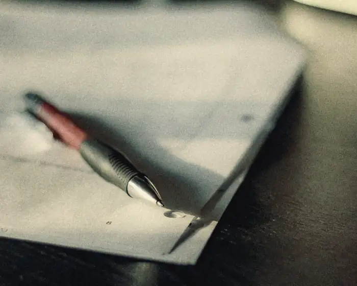 A pen resting on a stapled document on a dark desk, its shadow across the page